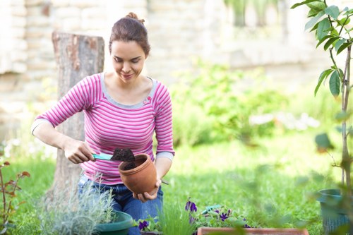 The image shows a young woman with dark hair tied back, wearing a pink and white striped long-sleeve shirt, tending to a garden in a suburban outdoor space. She is holding a small terracotta pot filled with soil in one hand and using a trowel with the other to transfer the soil. Her focus is directed downward towards the plants and soil, indicating active gardening work. The garden features a lush, green lawn with a variety of plants, including a small purple-flowered plant in a pot in the foreground, and other greenery and flowering plants nearby. In the background, there is a weathered wooden fence and a tree trunk, suggesting a well-maintained, private garden area. The lighting is natural, indicating a bright, possibly sunny day, with a softly blurred background that emphasizes the gardening activity. This scene reflects typical outdoor gardening tasks and highlights the importance of garden care provided by services like Lawn Mowing Stratford, situated within the Stratford area, showcasing a healthy, vibrant garden environment.