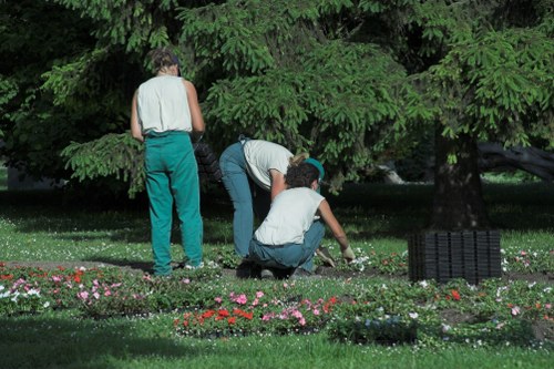 A garden scene featuring three individuals engaged in gardening activities beneath a large, leafy green tree. The foreground shows a flower bed with small, colorful flowering plants arranged along the edge of a lush, well-maintained lawn area. One person is kneeling on the grass, tending to the plants, while another is bent over, possibly planting or weeding, and the third stands nearby observing or assisting. The garden includes a mix of grass, soil, and flower borders, with the background dominated by dense foliage and tall trees, suggesting a peaceful outdoor space in Stratford. The weather appears to be clear and bright, highlighting the natural tones of the greenery and flowers. This scene reflects outdoor maintenance and gardening tasks typical of a professional landscaping or lawn care service, such as Lawn Mowing Stratford, aimed at maintaining a tidy and attractive garden environment.